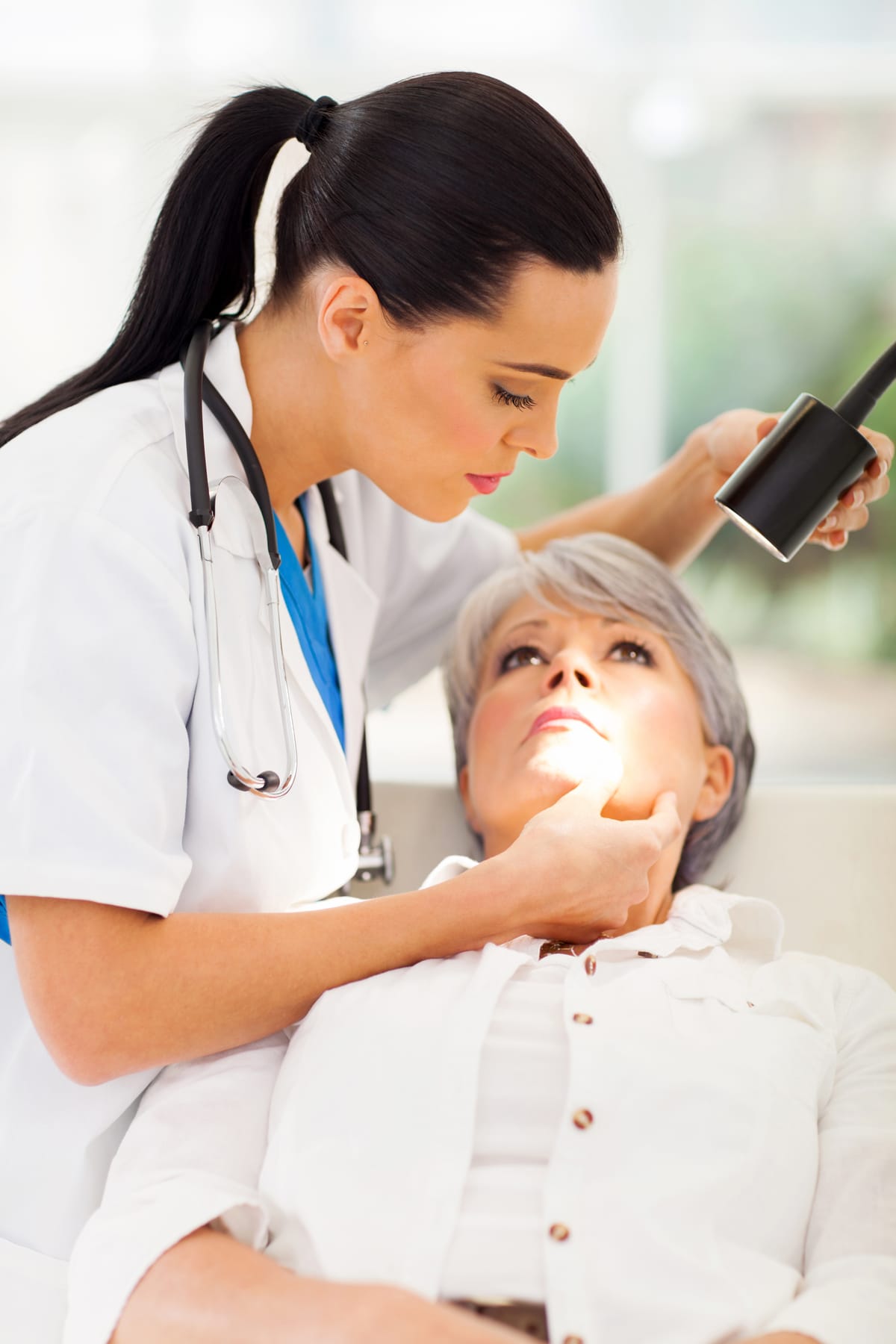 A dermatologist examining an older woman's facial skin with a lighted magnifier (models)