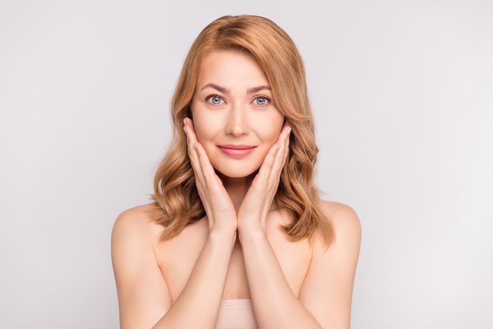 Middle-aged woman touching her smoother, shining face and smiling after receiving skin treatments.